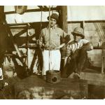 A photo of two International Drillers inside a rig with wooden siding. One is standing and holding a rope that leads into a dark box with a cylindrical top, while the other is sitting on a wooden board.