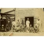 Six men standing and eight men sitting in front of a stone building with a set of wooden doors that are open.