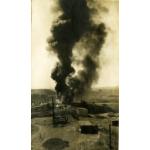 Thick dark smoke coming off of an oil well. There is a group of people off to the side and equipment in the foreground. There are hills in the background.