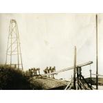 A photo of six men pushing down on a wooden crossbar supported by a wooden frame. There is a wooden four-pole structure behind them and a bush to the right.