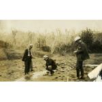 Four men looking down at a small water spring running through a bed of dirt and rocks. There are trees with no leave in the background.