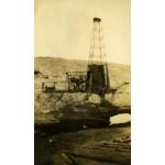 A metal oil rig. The base is covered in dark cloth. There is a roof over the wheels and belts beside the rig. There is casing on the ground in front of the rig and a bare hill in the background.