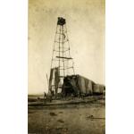 An oil derrick with four poles. The pole on the right is bent out of shape. There are three men on the top of the derrick and another climbing towards the, There is a group of men at the bottom of the rig, and part of the rig is covered in dark cloth.