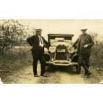 Webb and Burns standing on either side of a car parked on a dirt road. There are short trees on either side of them. The licence plate between the headlights reads: 1924. I.P.Co. STA.ELENA. 40".