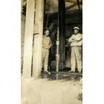 Two men wearing work clothes and hats standing inside a drilling rig. There is a large pipe and a wheel in behind them.