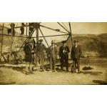 Five International Drillers standing at the base of a metal oil right, wearing dark suits and hats. There are men working on the lowest crossbeam. There are hills in the background.