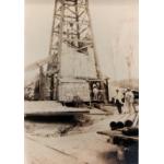 A photo of the bottom half of an oil rig being thatched. A group of men stand at the base. There are piles of casing on either side. 