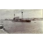 A ship with two masts and four smoke stacks anchored in the middle of a waterway. There are two large ships behind it and two small boats floating around it.