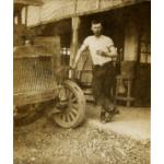 Ernest Kells wearing a white t-shirt and dark pants. He is resting his hand just above the wheel of a car and is standing in front of a building.