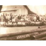 Cecil Coutts sitting in the middle of a local drilling crew in Burma. The group is sitting in front of a corrugated metal building, with casing on the ground in front.