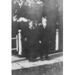 Winnett standing on a paved path outside their home. There is a white gate behind them and trees in front of the house.