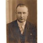 Portrait photo d'un homme portant un complet foncé et une cravate pâle. Ses cheveux sont lissés vers l’arrière et une toile de fond simple est derrière lui.