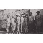 Seven men standing in front of a structure covered in metal siding. Eric Hussey stands in the middle with his arms around two men. He is wearing a pith helmet and coveralls.