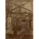 Two men standing in front of a large wheel with a belt attached to it, inside an oil rig. They are wearing white clothes and pith helmets. There are two workers behind them.