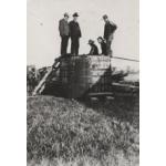 Un groupe de cinq hommes se tiennent sur le dessus d’un réservoir de stockage en bois de surface. Des anneaux de métal autour du baril le tiennent ensemble. Une échelle est appuyée contre le réservoir pour que les hommes puissent grimper sur celui-ci.