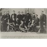 A group of men in front of a painted background. They are wearing dark jackets. Seven are standing, three are sitting in chairs, and one is sitting on the floor.
