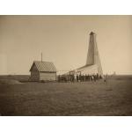 A group of people standing in front of an oil rig that has been covered in wooden planks. They are in a flat field and there is another rig in the background.