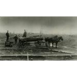 Two horses are harnessed to a cylindrical tank wagon with four wheels. Three men stand behind the wagon and there are oil rigs in the background.