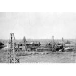 A field with three oil derricks in it. There are buildings in behind with pipes running between them. There are more derricks in the background. Along the bottom of the photo, "Woodward's Refinery" is written in white.