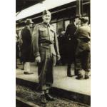 A man wearing a military uniform, standing beside railway tracks in front of a train. There are people standing on the platform behind him.