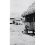 A man sitting on the steps leading up to a thatched building on stilts. There is another building in the background.