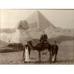 John Growder assit sur un chameau devant le Sphinx et une pyramide. Il y a un homme tenant les rênes du chameau et un autre à l'arrière du celui-ci.