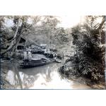 Six hommes dans un bateau sur une rivière étroite. Deux hommes sont sur un quai avec une pile de tubage à leurs côtés sur la rive. Un groupe d’hommes se tiennent derrière le tubage devant les trois bâtiments. Il y a des arbres en arrière-plan.