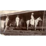 Deux hommes et une femme à cheval devant un bâtiment avec une terrasse surélevée. Les hommes montent des chevaux foncés et portent des chemises et des pantalons blancs. La femme est vêtue d'une robe blanche et d'un chapeau. Elle monte un cheval blanc.