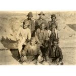 Charles Warren stands with seven local men arranged in three rows, all wearing work clothes. Warren is in the centre in the back row. The middle row is sitting on chairs and the front row is sitting cross-legged on the ground.