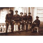 A photo of six men dressed in army uniforms and holding canes. They are sitting on a white railing in front of a building with white siding. The man on the right is sitting on a wicker chair.