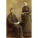 A photo of Charles and Jessie Nicklos. Charles is sitting in a chair and holding a piece of paper while Jessie is standing with her arms folded.