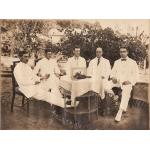 Five men sit around a wicker table covered with a white tablecloth. There is a bouquet of flowers in front of a frame on the table. The men are wearing white suits.