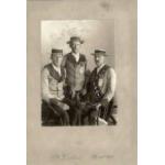 Three men wearing jackets and boat hats. The man in the middle is standing and the other two are sitting on chairs. The man on the right is wearing a sash.