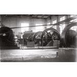 Inside a refinery with four large wheels attached to smaller wheels with belts of fabric. Men stand near the wheels. There are windows letting light into the room.