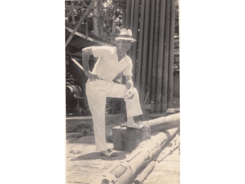 Harry Smith dressed in a white suit and panama hat, with one leg up on a bench.