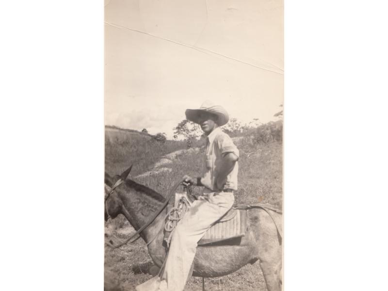 Harry Smith with a big hat, sitting on a mule.  He is wearing white and there are hills in the background.