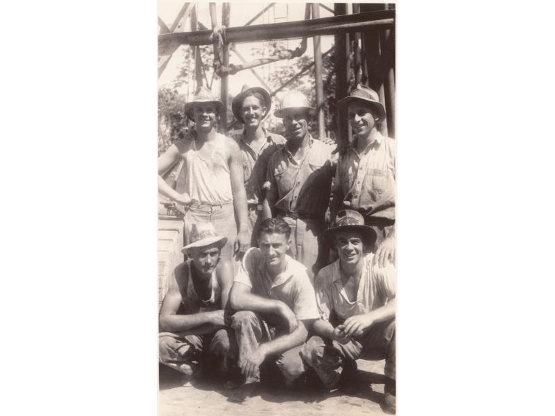 Un groupe de sept foreurs internationaux sous une structure de métal. Quatre sont debout et trois à genoux. Ils portent des vêtements de travail.