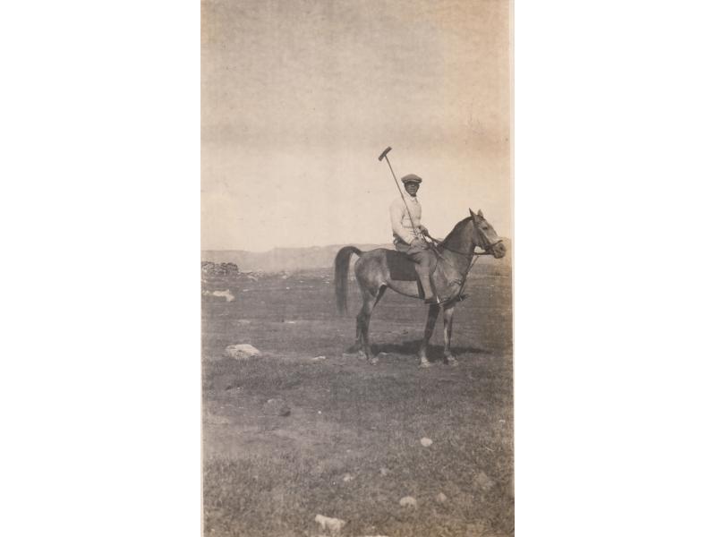 Harry Smith riding a horse in a stony field, holding a polo stick. There are hills in the background.