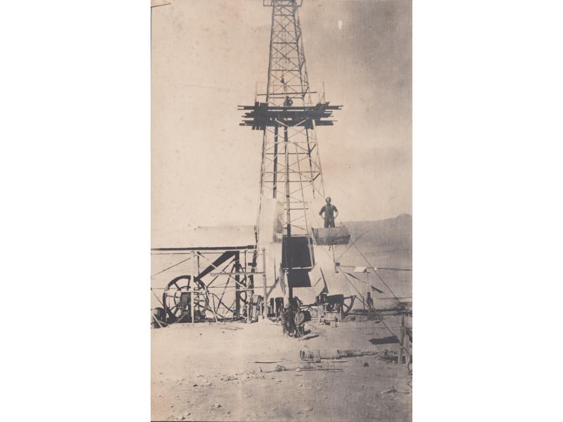 A small group of people standing at the bottom of an exposed oil rig. There is a man standing in a bucket suspended by lines above them.