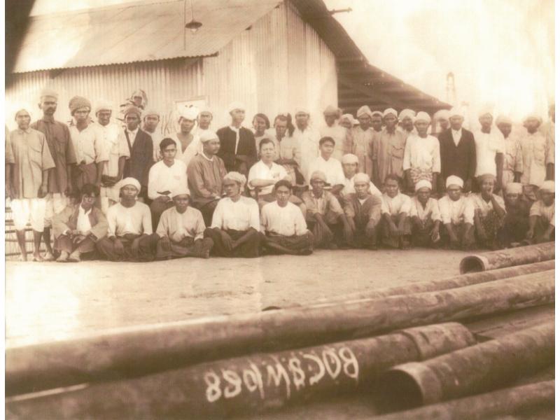 Cecil Coutts sitting in the middle of a local drilling crew in Burma. The group is sitting in front of a corrugated metal building, with casing on the ground in front.