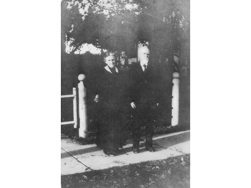 Winnett standing on a paved path outside their home. There is a white gate behind them and trees in front of the house.