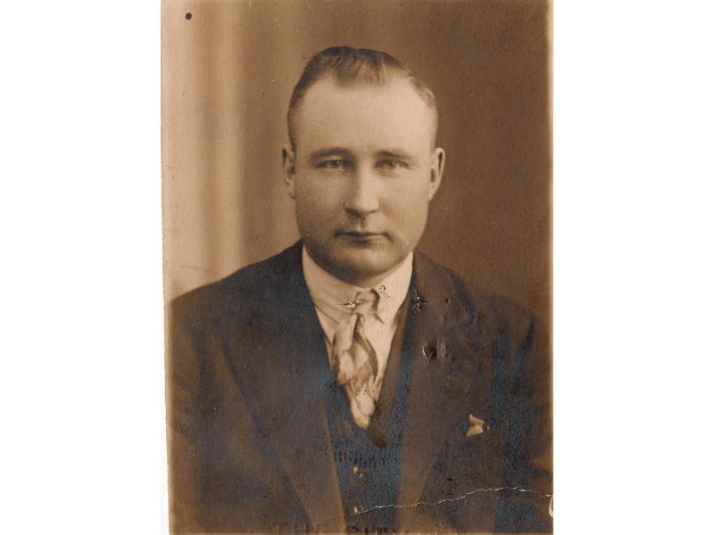 A portrait photo of a man wearing a dark suite and light tie. His hair is slicked back and there is a plain background behind him.