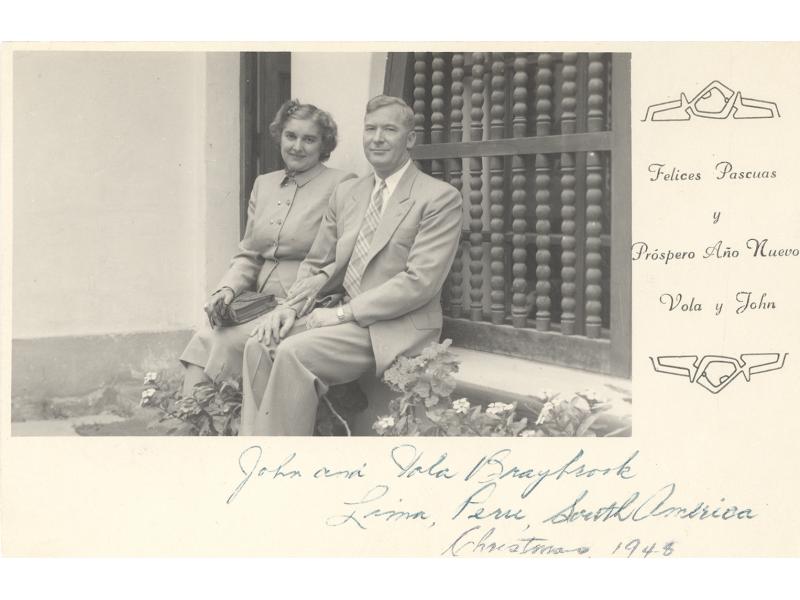 Vola and John Braybrook sitting on a windowsill. There are bars on the window and flowers by their feet. The photo is part of a Christmas card.