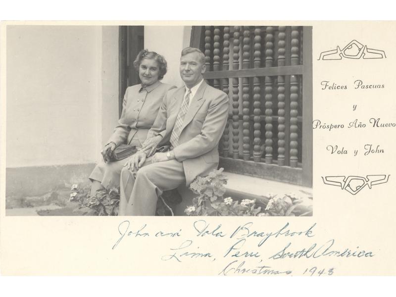 Vola et John Braybrook assis sur le bord d’une fenêtre. Il y a des barreaux à la fenêtre et des fleurs à leurs pieds. La photo fait partie d’une carte de Noël.