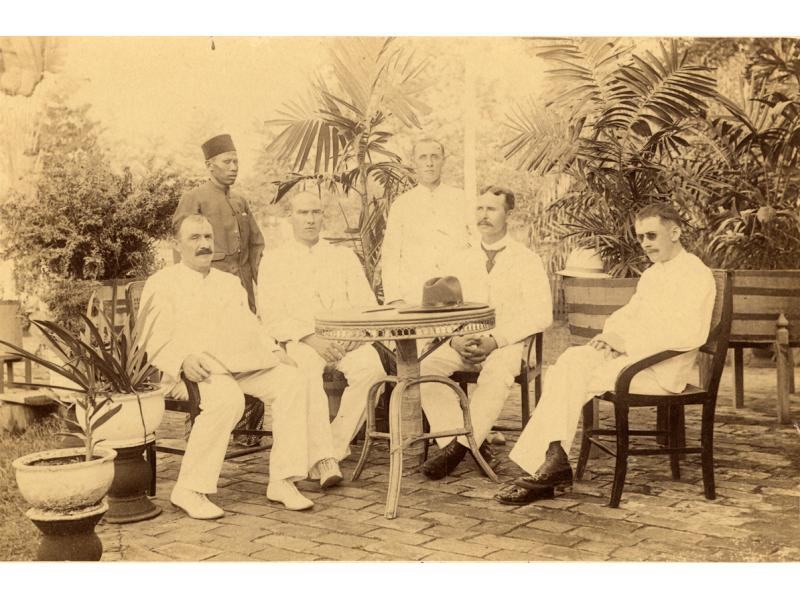 A photo of five International Drillers, wearing white, around a table with a hat on it. A servant stands behind them. There are trees in the background.