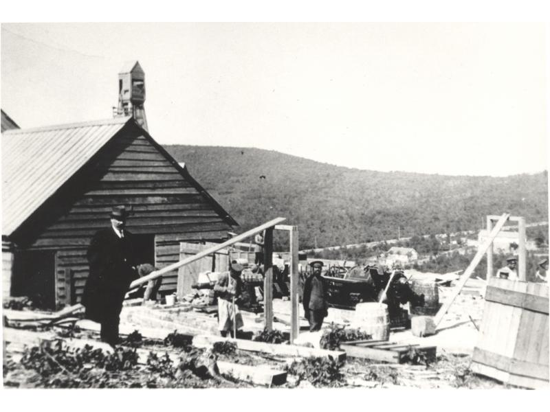 Charles Wallen debout dans un champ de pétrole devant un bâtiment en bois. Des travailleurs et de la machinerie se trouvent derrière lui. On aperçoit une colline couverte d'arbres en arrière-plan.