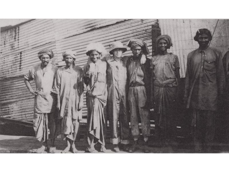Seven men standing in front of a structure covered in metal siding. Eric Hussey stands in the middle with his arms around two men. He is wearing a pith helmet and coveralls.