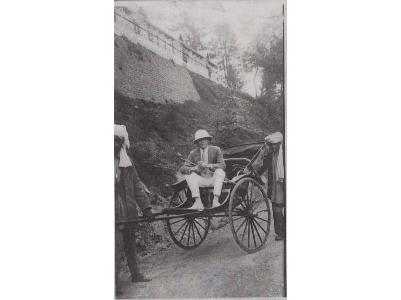 Eric Hussey sitting in a rickshaw being pulled by a man. Another man stands beside the rickshaw. They are on a road at the bottom of a hill with a building on top of it.