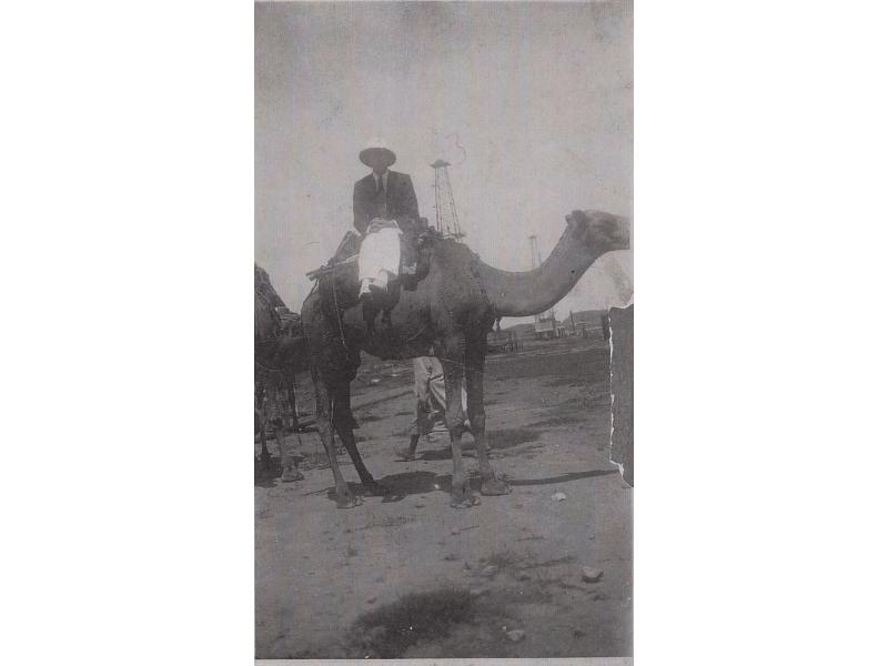 Eric Hussey sitting sideways on a camel, facing the camera. He is wearing a pith helmet. There are two oil derricks in the background.