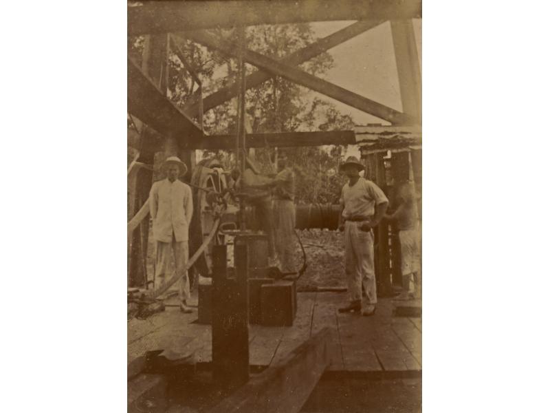 Two men standing in front of a large wheel with a belt attached to it, inside an oil rig. They are wearing white clothes and pith helmets. There are two workers behind them.